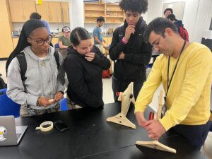 A Physics teacher stands by a catapult with three students.