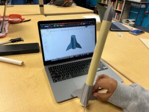 A student holds up a rocket next to a laptop screen with the computer assisted drawing of the fins of the rocket.